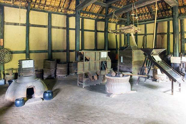 A hearth, wooden containers, a large mortar and other household or work objects displayed together beneath exposed wooden beams.
