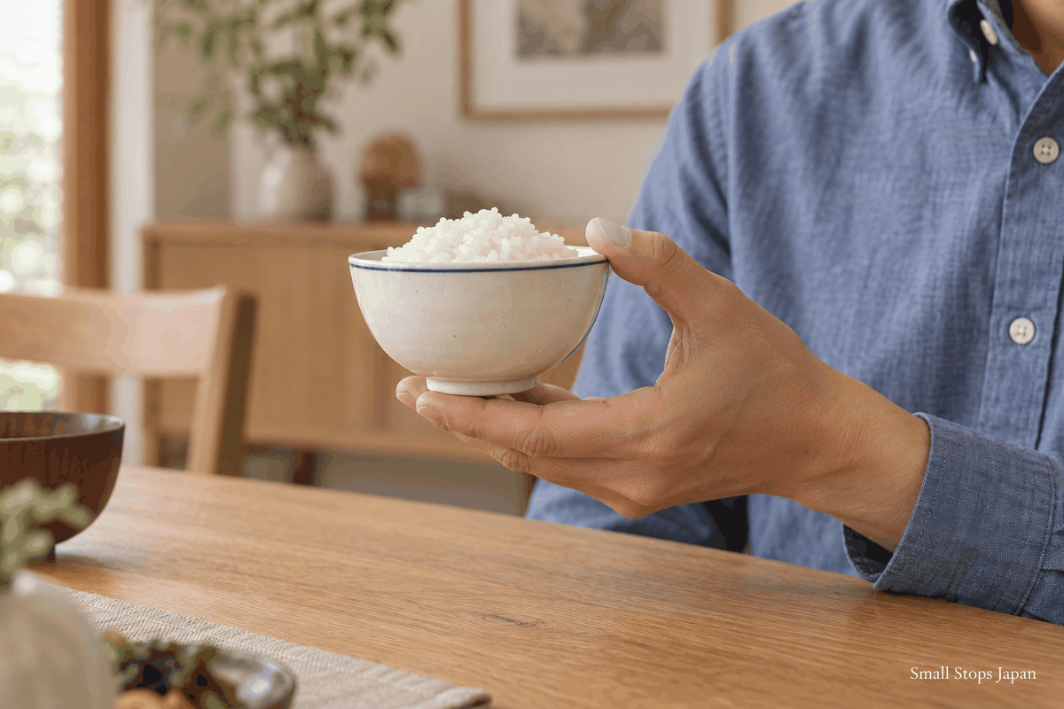 An illustrated left hand with fingers together under a rice bowl's foot ring and the thumb lightly at its outer rim.