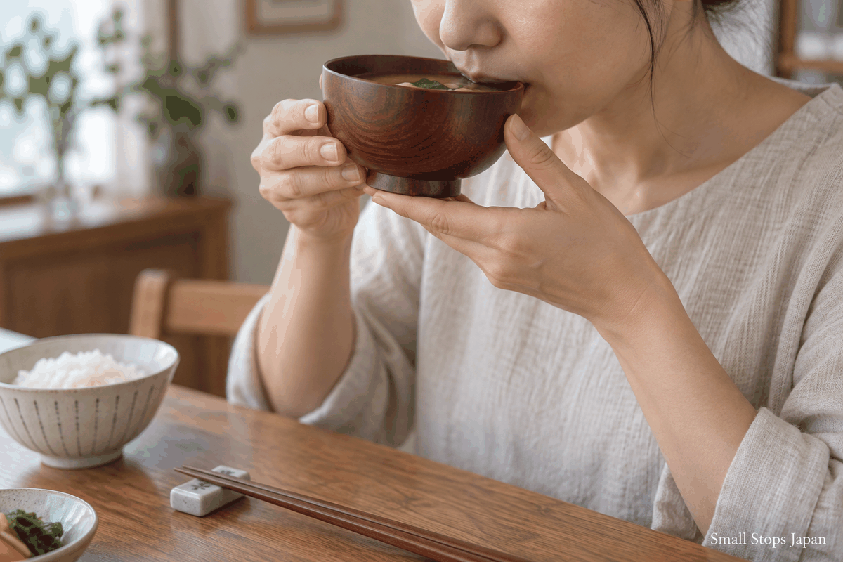An adult sipping from a small soup bowl, supporting its base and steadying its side while chopsticks rest on the table.