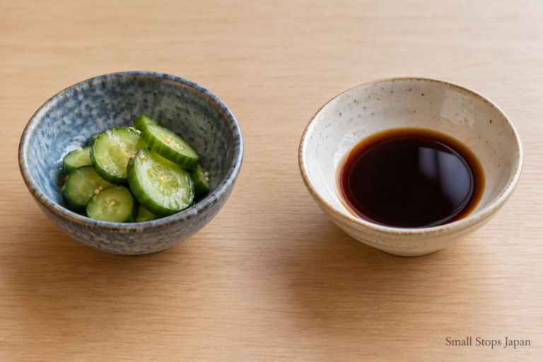 Two separate small bowls, one holding sliced cucumber and the other a shallow pool of soy sauce.