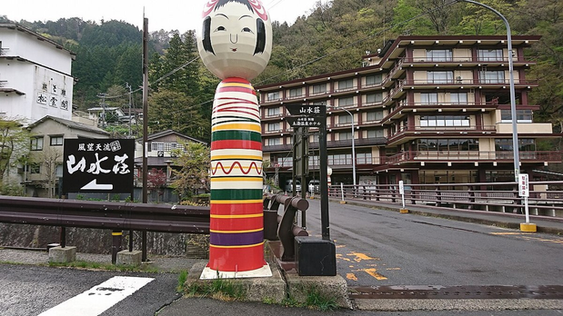 A giant painted kokeshi beside a bridge, with buildings and wooded hills in Tsuchiyu Onsen behind it.