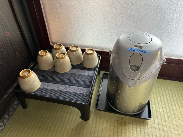 Six inverted ceramic cups on a low stand beside a dispenser labelled cold barley tea.