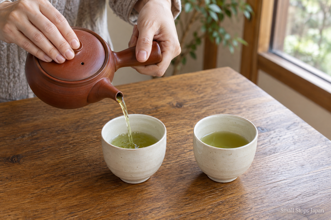 Tea pours from a side-handled teapot into one of two pale-interior cups, with the tea visible in the other.