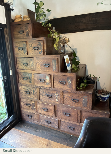 Stepped wooden storage with many drawers at Bunke Tamaya, with plants, a framed picture and a black telephone on the steps.