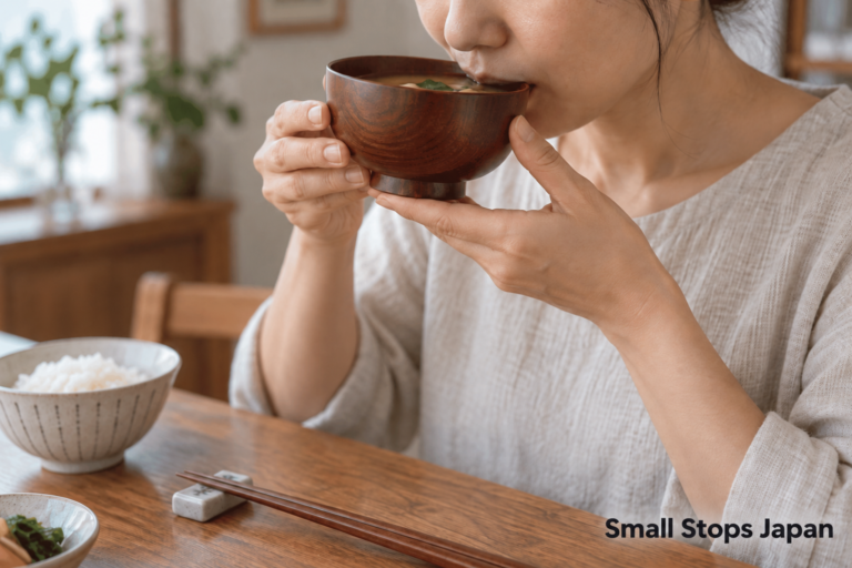 An adult holds a soup bowl at the mouth with both hands while chopsticks lie on a rest.