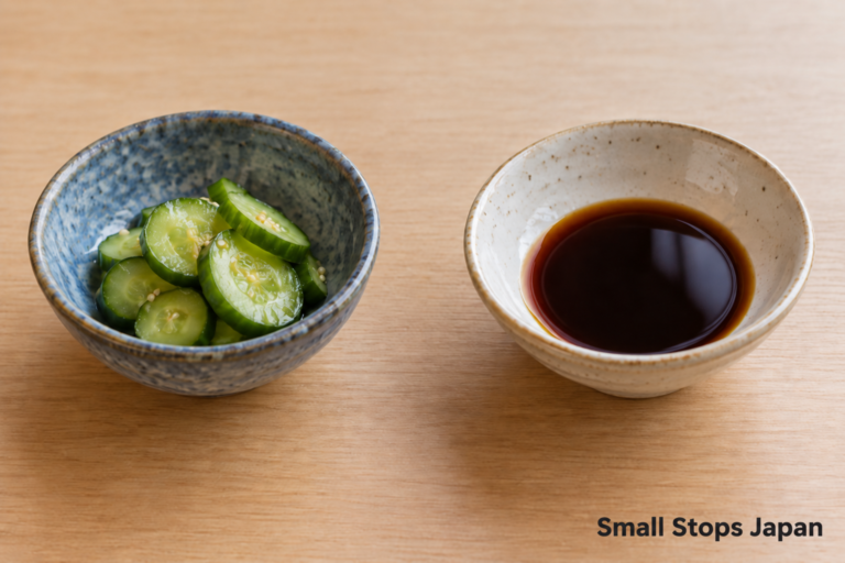 Two small bowls, one with sliced cucumber and one with soy sauce.