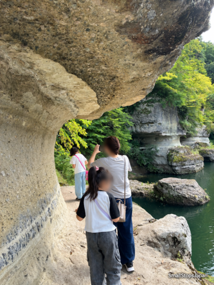 An overhanging rock face above a narrow, uneven riverside passage, with water and rocks beside it.